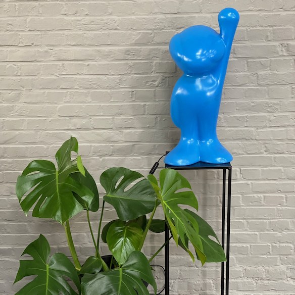Eine leuchtend blaue Figur über grünen Blättern vor einer hellen Backsteinwand