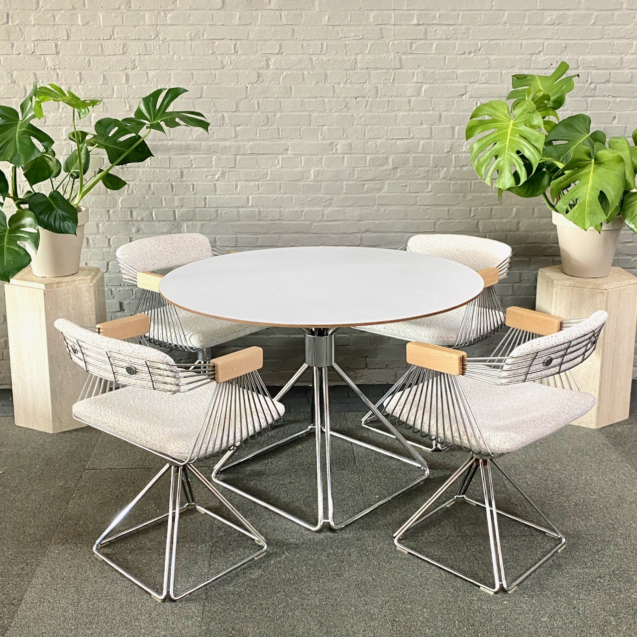 A pale round dining table with wire-frame chairs and green plants against a brick wall