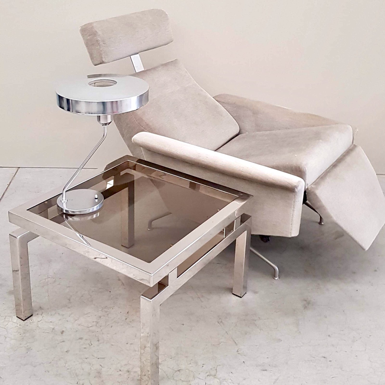 Een ivoorkleurige fauteuil met een verchroomde tafellamp en een lage tafel met glazen blad